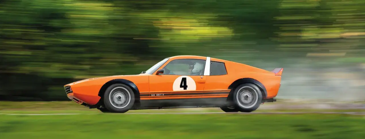 En orange Saab Sonett IV med nummer 4 på sidan susar fram på en bana, omgiven av suddiga gröna träd.