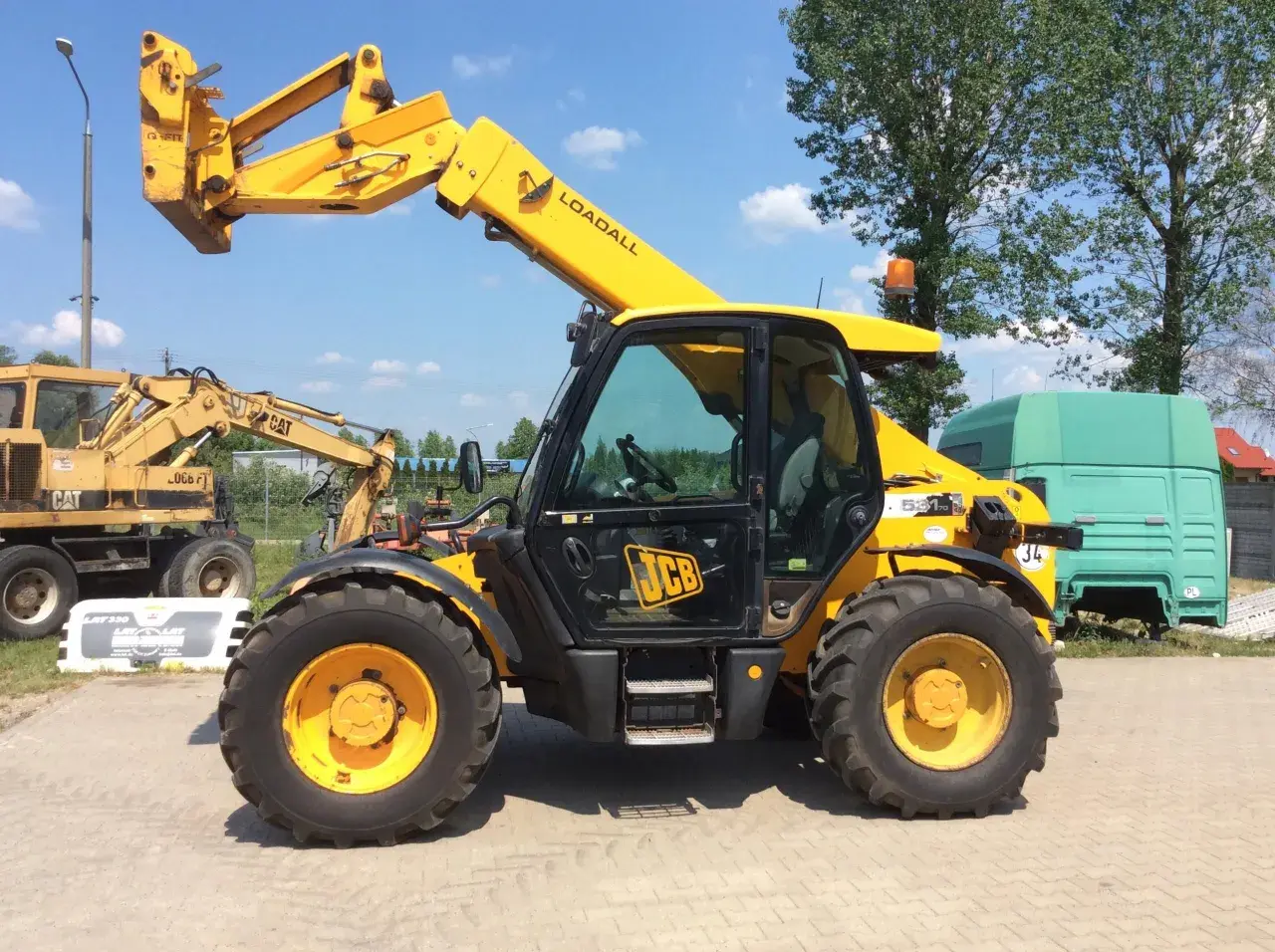 Żółty ładowarka teleskopowa JCB 531-70 Loadall stoi na placu. W tle koparka CAT i zielona kabina ciężarówki.