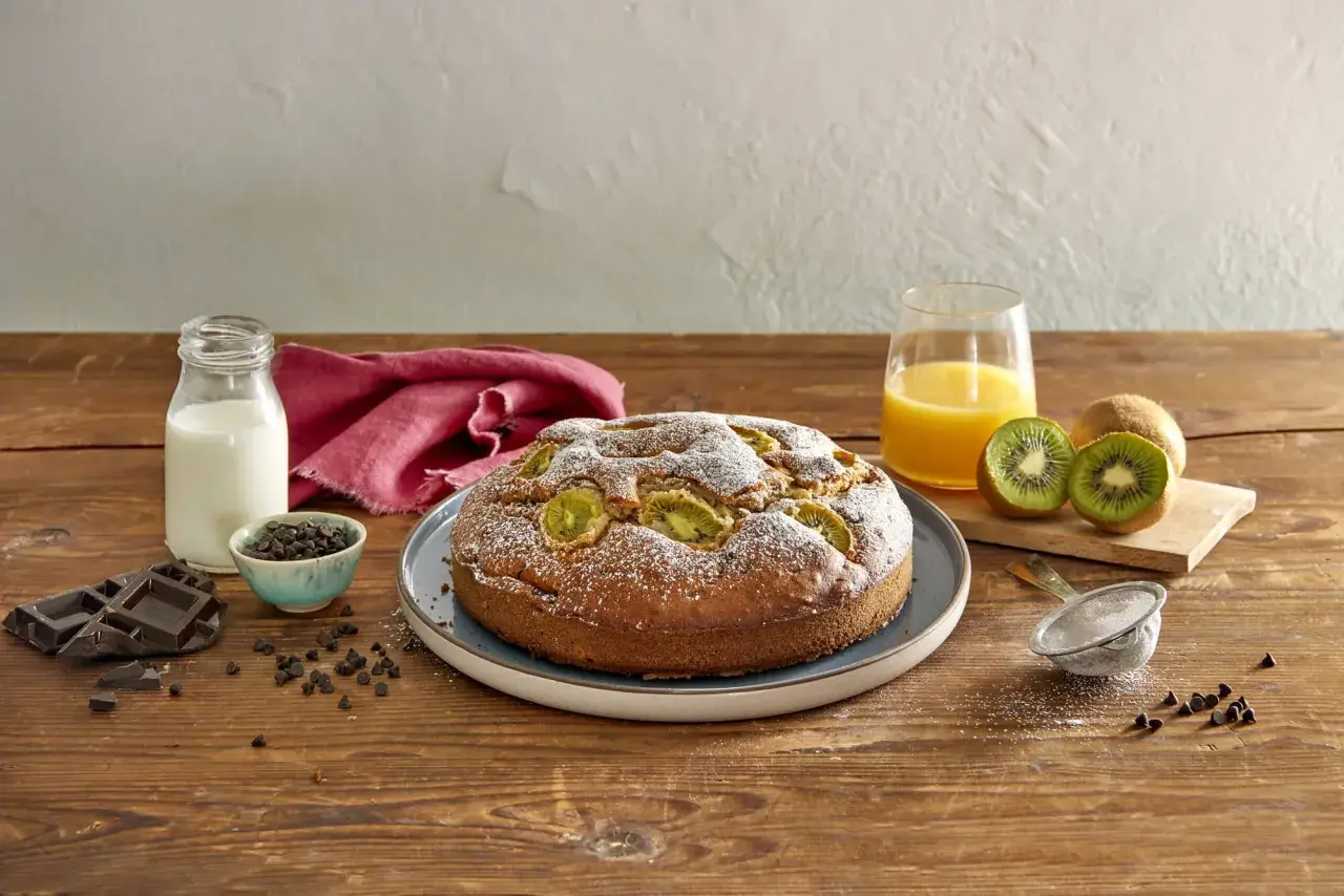 Torta kiwi e cioccolato cosparsa di zucchero a velo, con kiwi freschi, gocce di cioccolato, latte e succo d'arancia su un tavolo di legno.