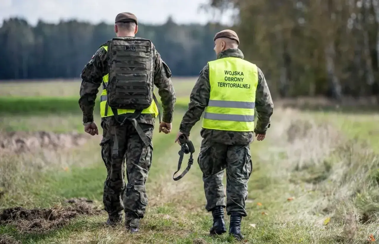 Dwóch żołnierzy Wojsk Obrony Terytorialnej w kamizelkach odblaskowych idzie przez pole. Rezygnacja z WOT może mieć poważne konsekwencje.