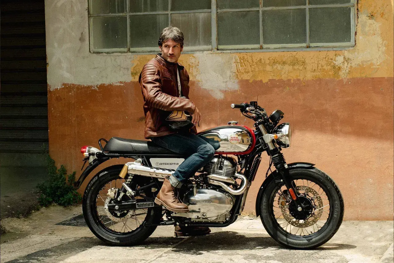 Uomo con giacca in pelle marrone seduto su una moto Royal Enfield Interceptor. Il manubrio cafe racer è cromato, pronto per un viaggio.