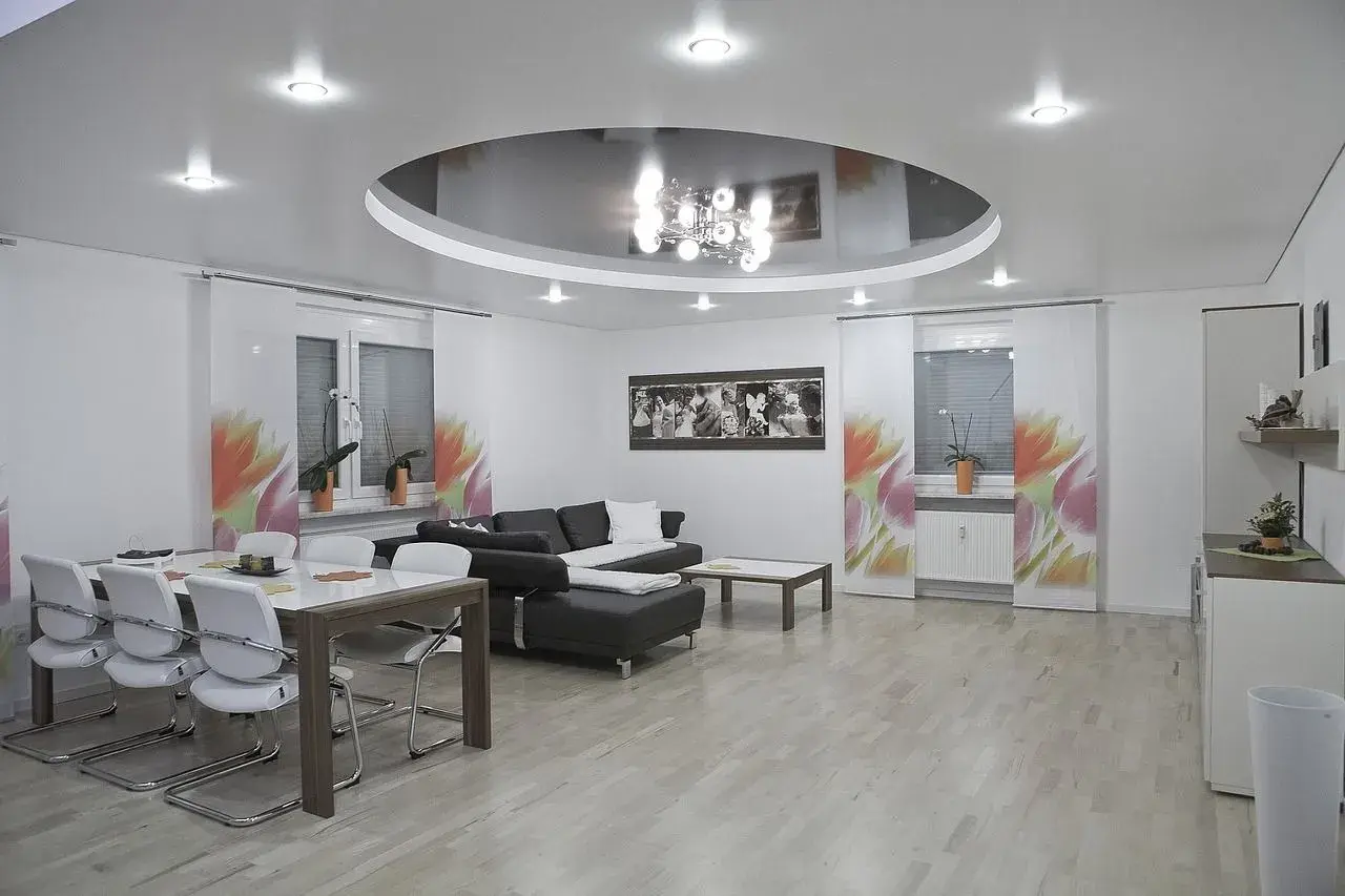 Nowoczesny salon z okrągłym sufitem napinanym, który odbija elegancki żyrandol. Wygodna kanapa i stół z krzesłami tworzą przytulną przestrzeń.