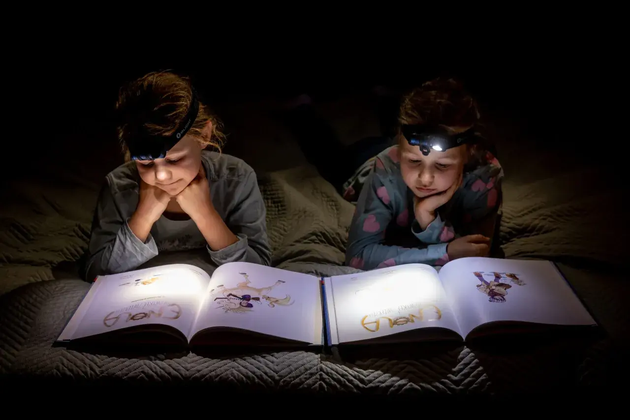 Dwie dziewczynki z latarkami na głowach czytają książkę w ciemności. To świetny sposób, by przygotować się na blackout i nie nudzić się!