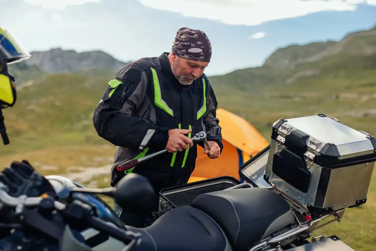 Hombre con chaqueta de moto repara su vehículo, quizás para evitar que la moto se ahogue. Montañas y tienda de campaña al fondo.