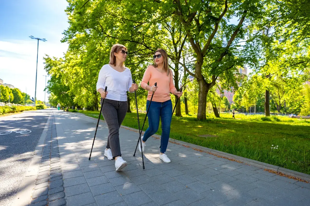 Deux femmes font de la marche nordique dans un parc ensoleillé, une activité idéale pour perdre du poids en marchant.