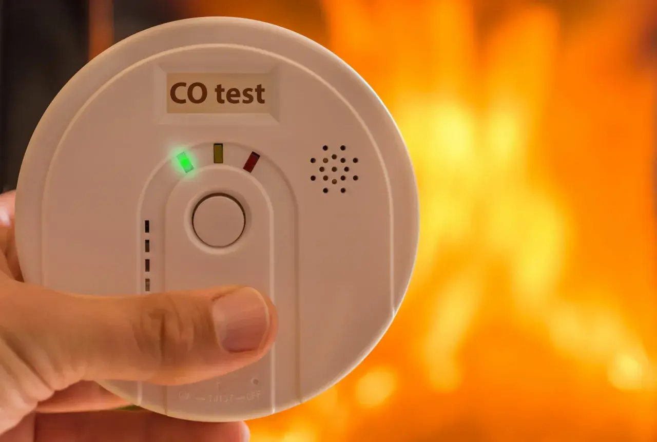 Détecteur de monoxyde de carbone tenu à la main, avec un feu de cheminée en arrière-plan. Où placer détecteur monoxyde de carbone ?