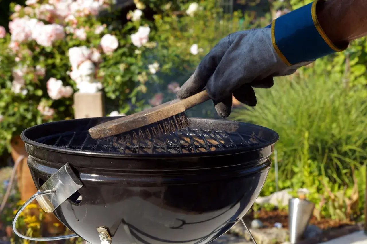 Mit einem Handschuh wird der Grillrost gereinigt. Rauch steigt auf, während die Bürste über die Roste fährt.