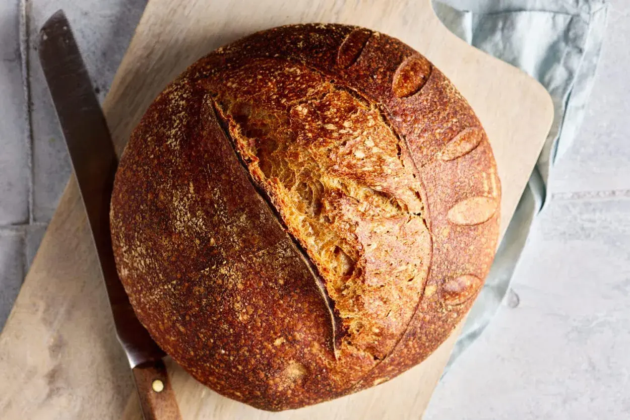 Ein rustikales Dinkel Sauerteigbrot mit knuspriger Kruste und luftiger Krume liegt auf einem Holzbrett neben einem Messer.