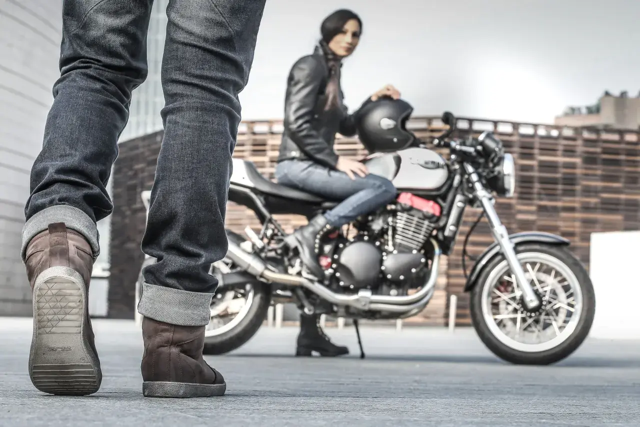 Kobieta w skórzanej kurtce siedzi na motocyklu, obok stoją nogi w brązowych butach motocyklowych.