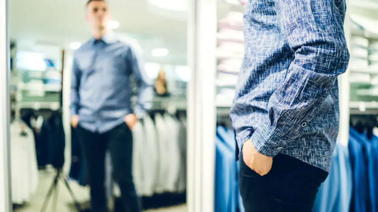 Mann im Spiegel prüft seinen lässigen kleidungsstil herren. Blaues Hemd mit Muster, schwarze Hose.