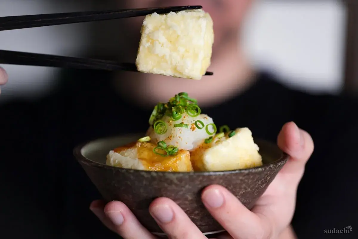 Chopsticks lift a piece of crispy age tofu, ready to be dipped into a savory sauce.