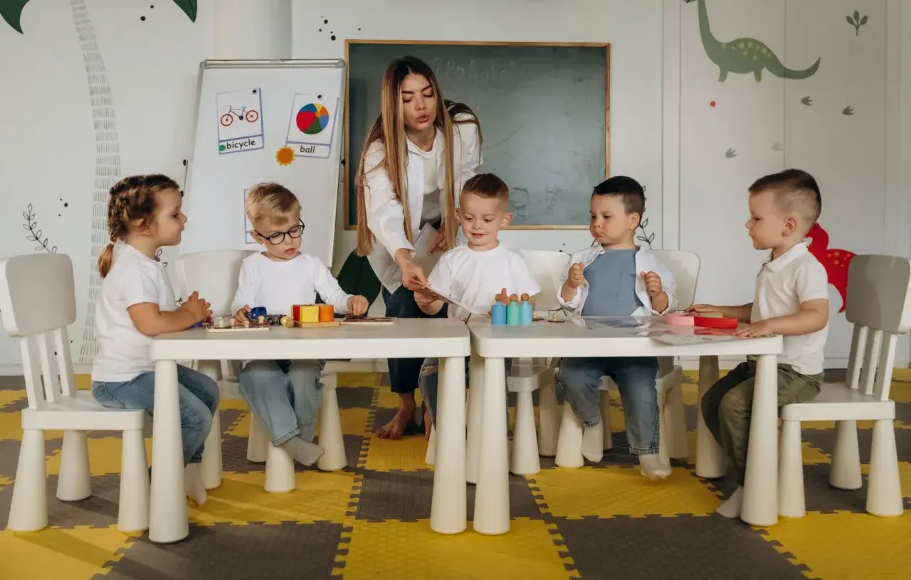 Nauczycielka bez wykształcenia pedagogicznego prowadzi zajęcia z dziećmi w przedszkolu. Dzieci siedzą przy stolikach, bawiąc się klockami i kartami.