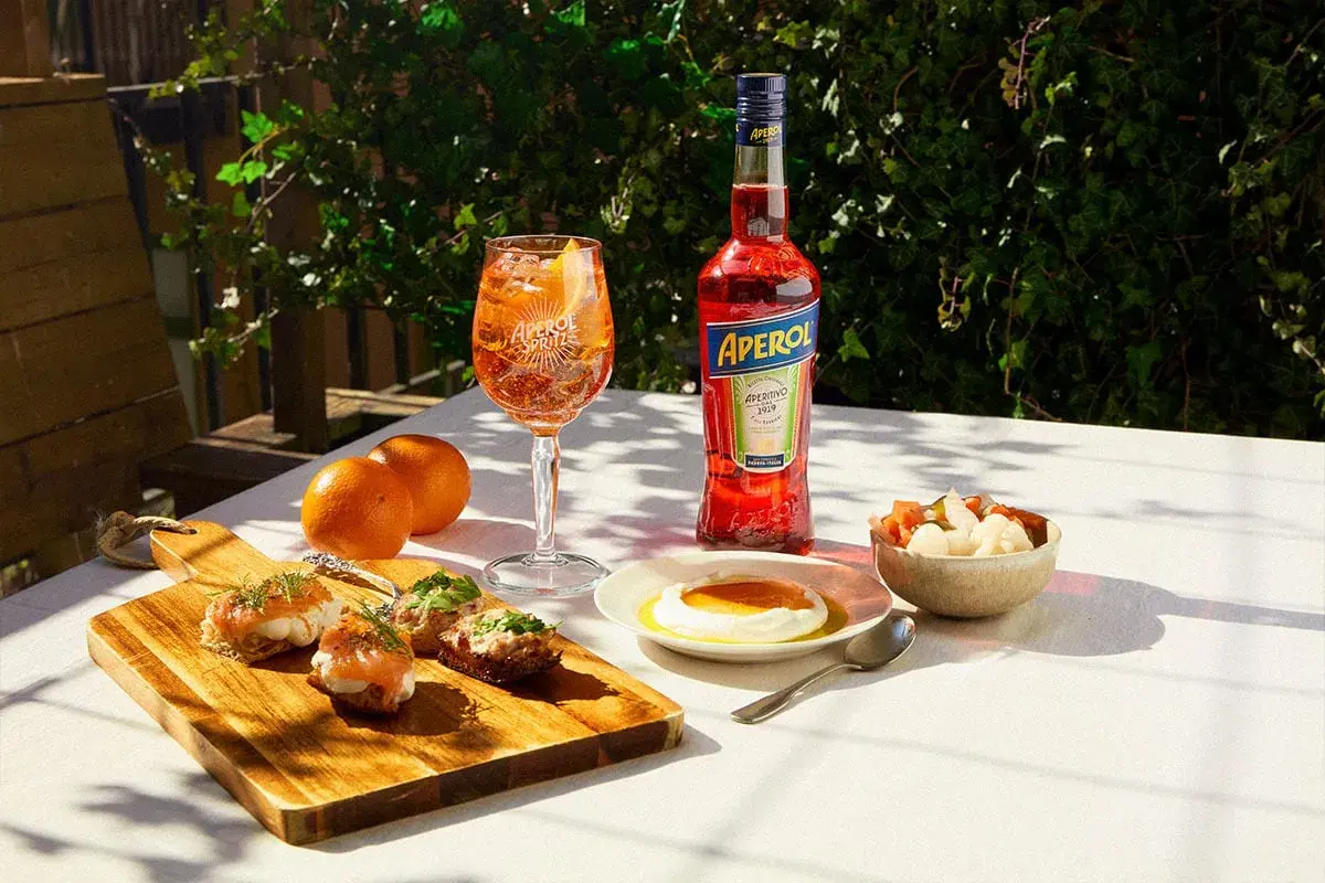En Aperol Spritz med tilltugg till aperol spritz: laxsnittar, ost med honung och inlagda grönsaker.