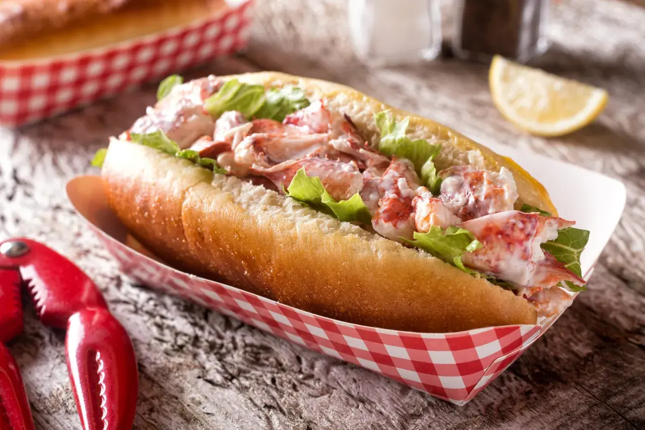 Pyszny lobster roll z kawałkami homara i sałatą w bułce, podany w czerwono-białym papierowym opakowaniu. Obok szczypce do homara.