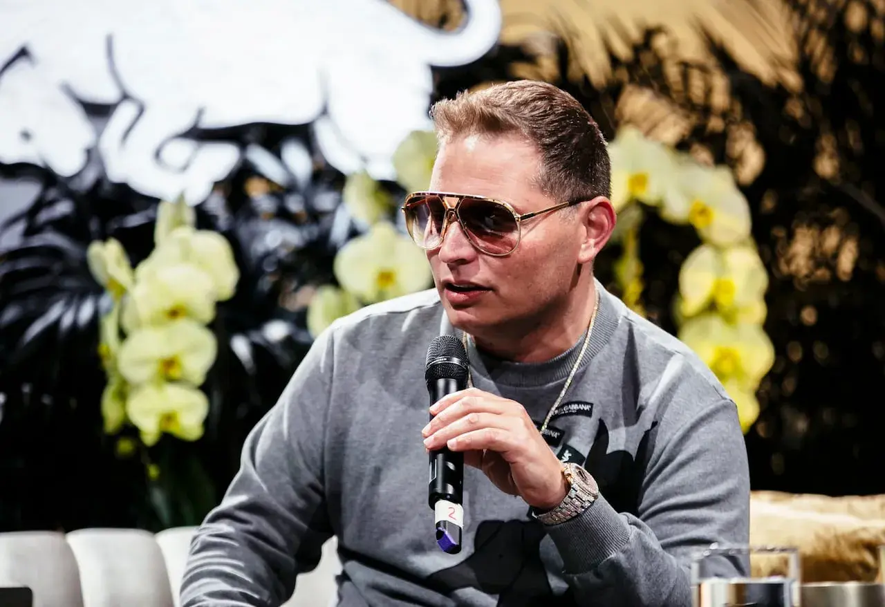 Scott Storch, known for his hit songs, speaks into a microphone, wearing sunglasses and a grey sweatshirt.