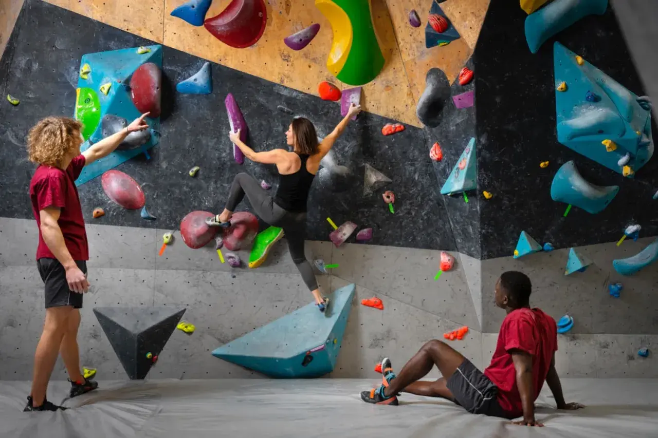 Drei Personen bouldern an einer bunten Wand. Ein Mann gibt Tipps, eine Frau klettert, ein Mann ruht.