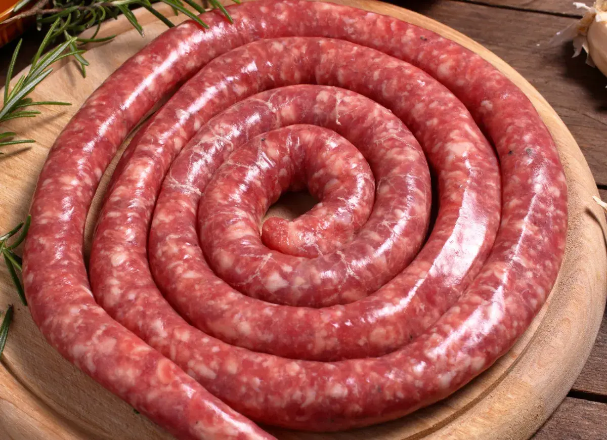 Surowa, zwinięta w spiralę salsiccia na drewnianej desce, ozdobiona gałązkami rozmarynu.