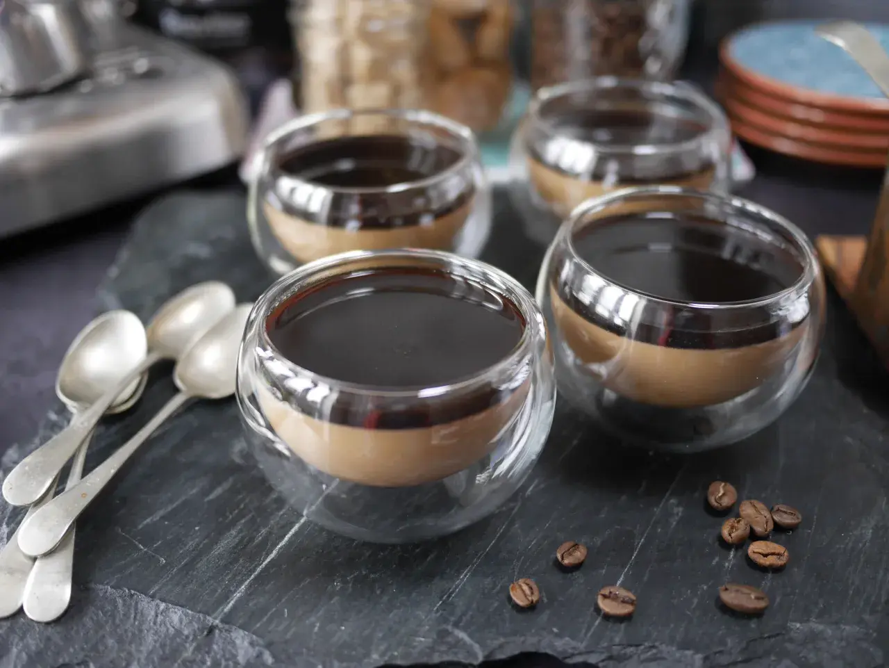 Fyra små glas med kaffe pannacotta, toppad med mörk sirap. Tre skedar ligger till vänster, och kaffebönor sprider sig på stenen.