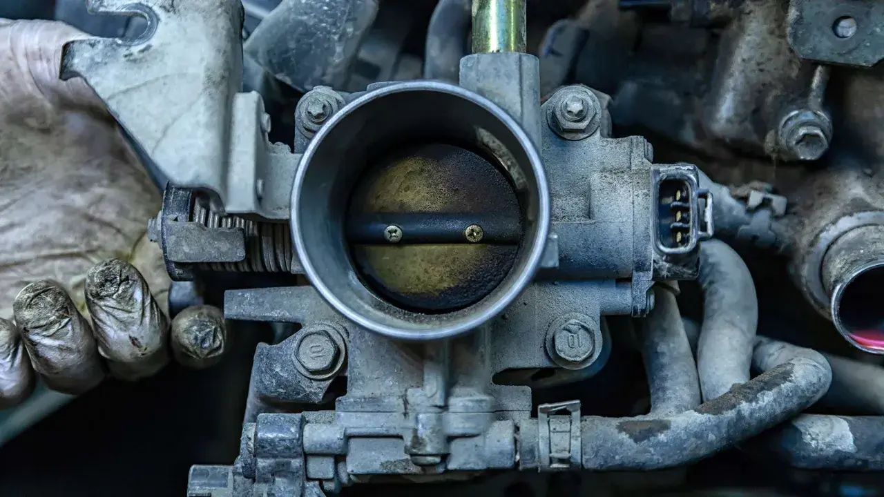 Primer plano de una mariposa de admisión de motor de coche, con suciedad y grasa. Unas manos enguantadas se ven a la izquierda.