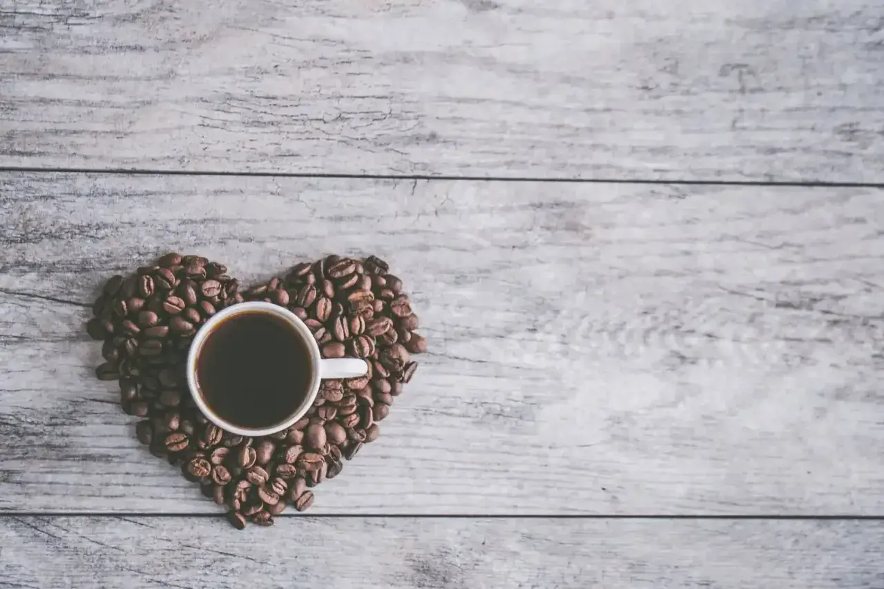 Una tazzina di caffè nero, circondata da chicchi di caffè disposti a forma di cuore su un tavolo di legno. Un invito al caffè in allattamento.