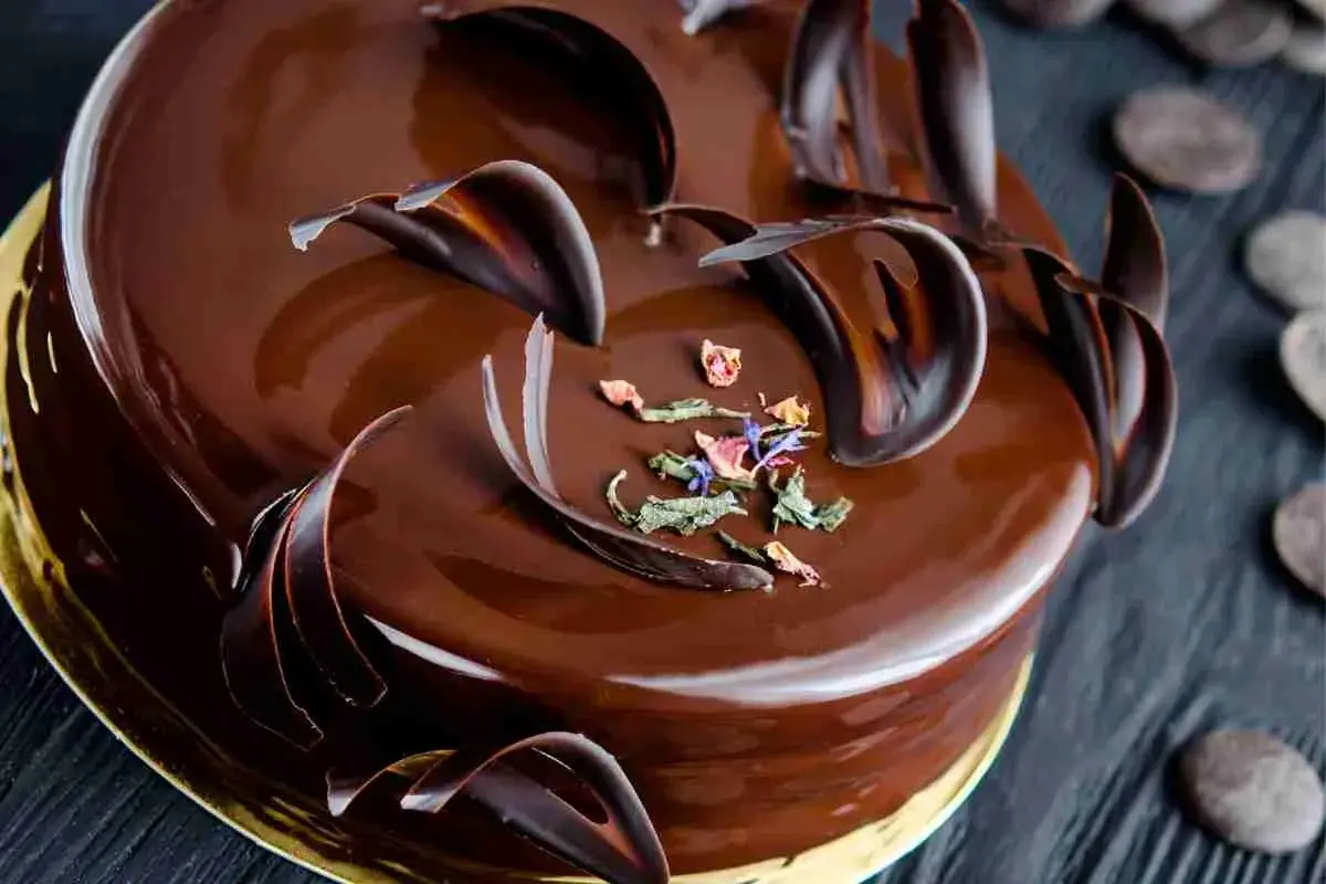 Una lucida torta Savoia al cioccolato, decorata con riccioli di cioccolato fondente e petali di fiori secchi.