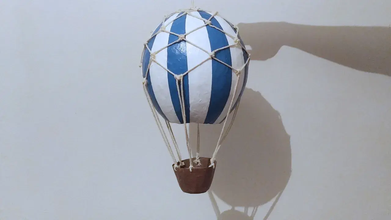 Globo aerostático manualidad, con rayas azules y blancas, suspendido con cuerdas y una cesta de mimbre.