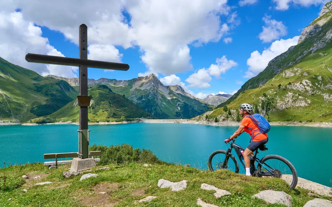 Ciclista en el Alpe Adria Radweg, junto a un lago turquesa y montañas verdes.