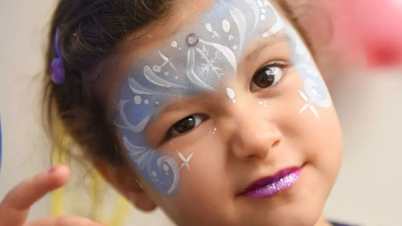 Bambina con trucco da fata del ghiaccio per il carnevale, con glitter viola sulle labbra e disegni bianchi e azzurri sul viso.