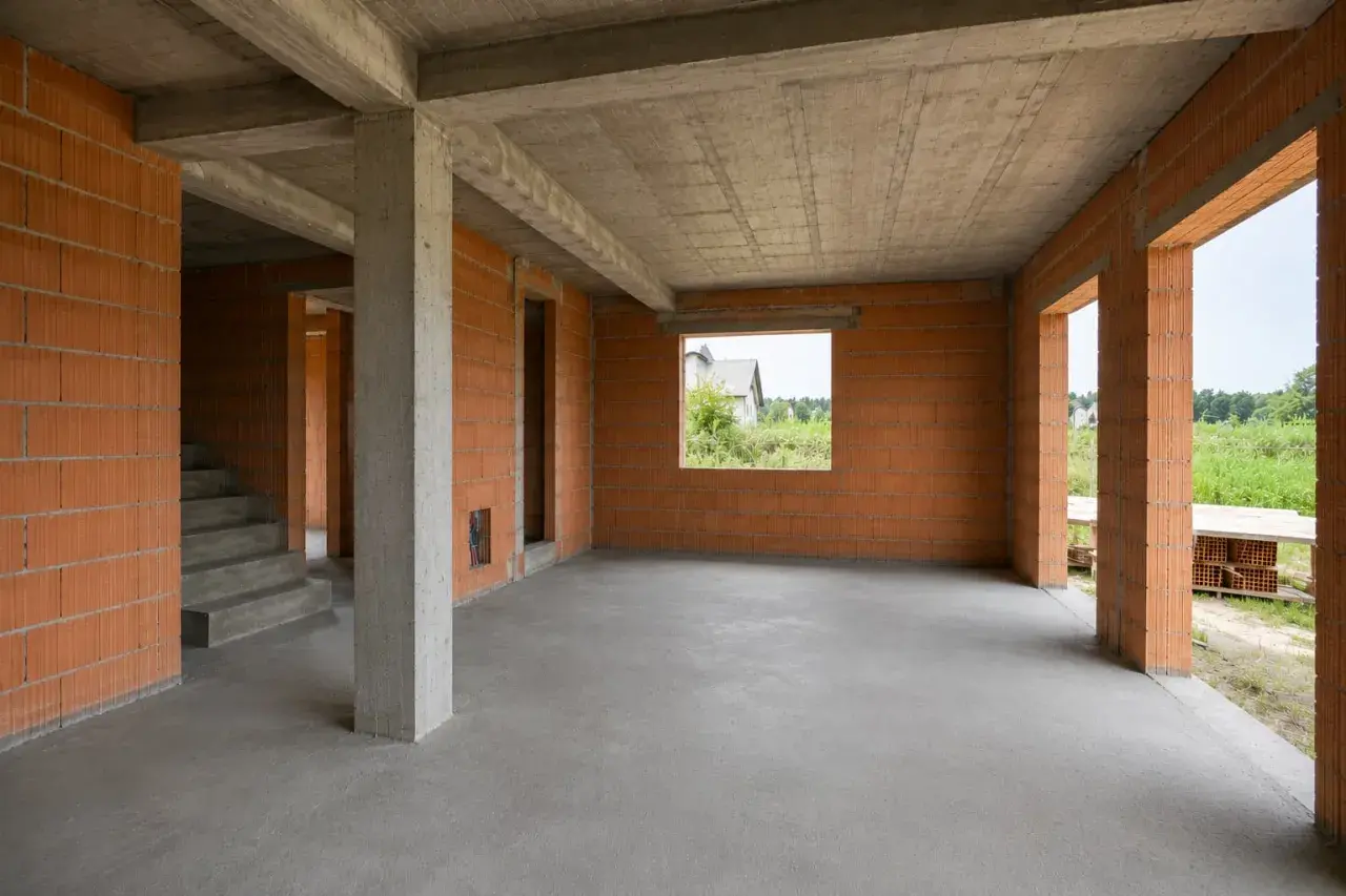 Wnętrze domu w stanie surowym z ceglanymi ścianami, betonowymi stropami i schodami.