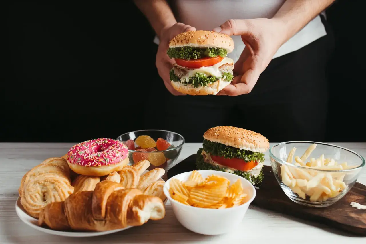 Ręce trzymają burgera, obok talerz z croissantami, pączkiem i ciastkami, miseczki z żelkami, frytkami i chipsami. Czy cheat day przyspiesza metabolizm?
