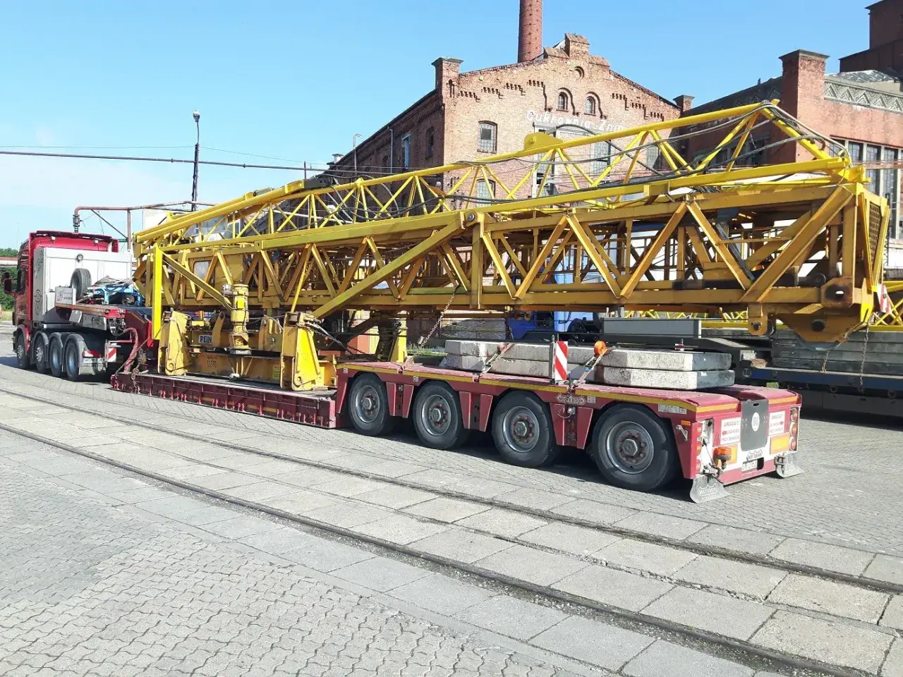 Ciężarówka z przyczepą transportuje żółty element dźwigu. Wymaga to zezwolenia na transport ponadgabarytowy.