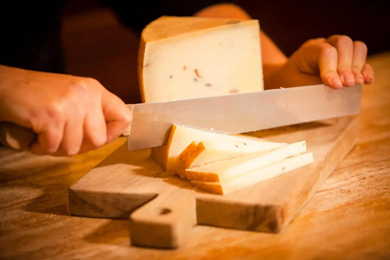 Mani che tagliano formaggio su un tagliere di legno. Fette sottili e un pezzo più grande di formaggio stagionato.