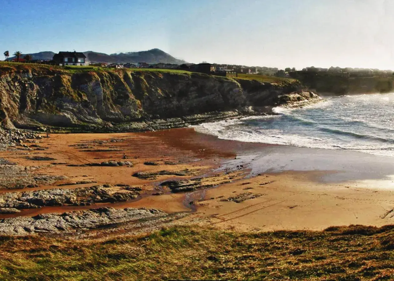 La playa de Portio, con sus acantilados rocosos y arena dorada, se baña en la luz del sol. Las olas rompen suavemente en la costa.
