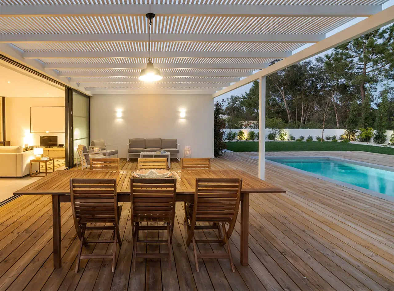 Table et chaises en bois huilé sur une terrasse en bois, avec piscine et jardin en arrière-plan.