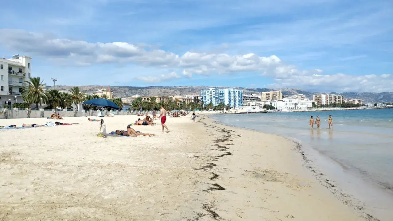 Spiaggia di Manfredonia con persone che prendono il sole e tre donne che camminano nell'acqua cristallina.