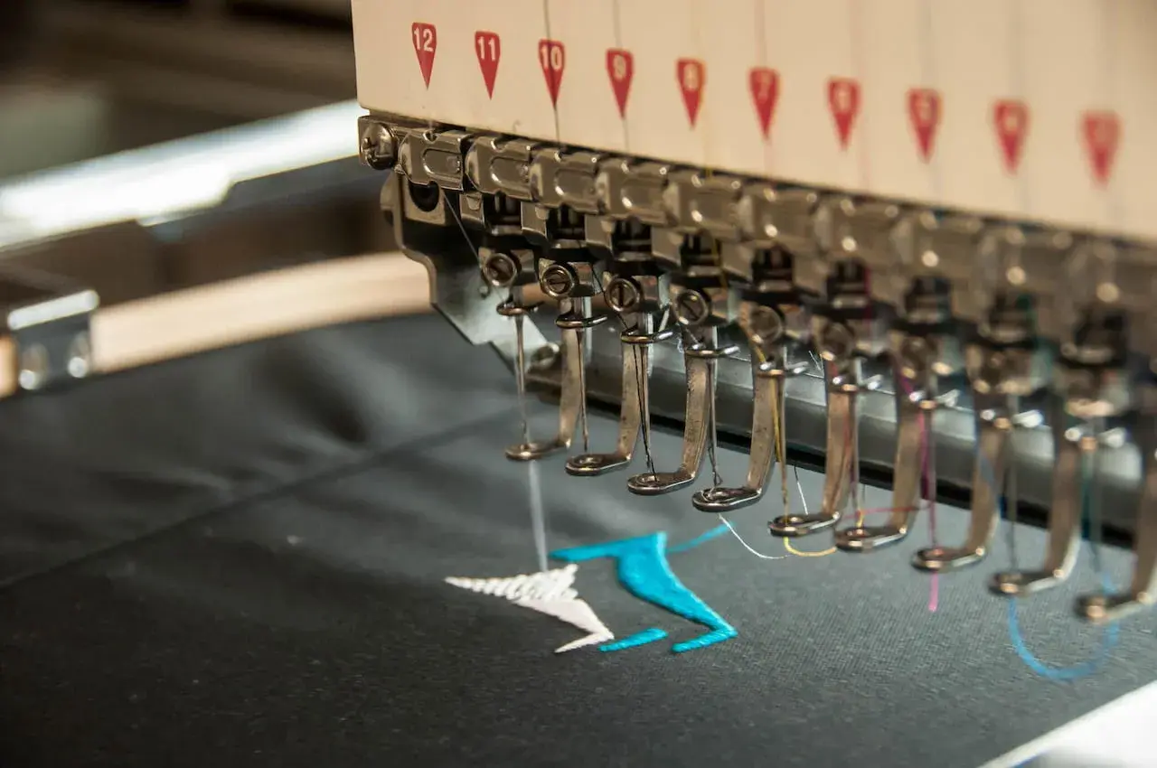 Machine à broder en action, créant un motif coloré sur un tissu noir. La **direct to garment printing** permet des personnalisations uniques.