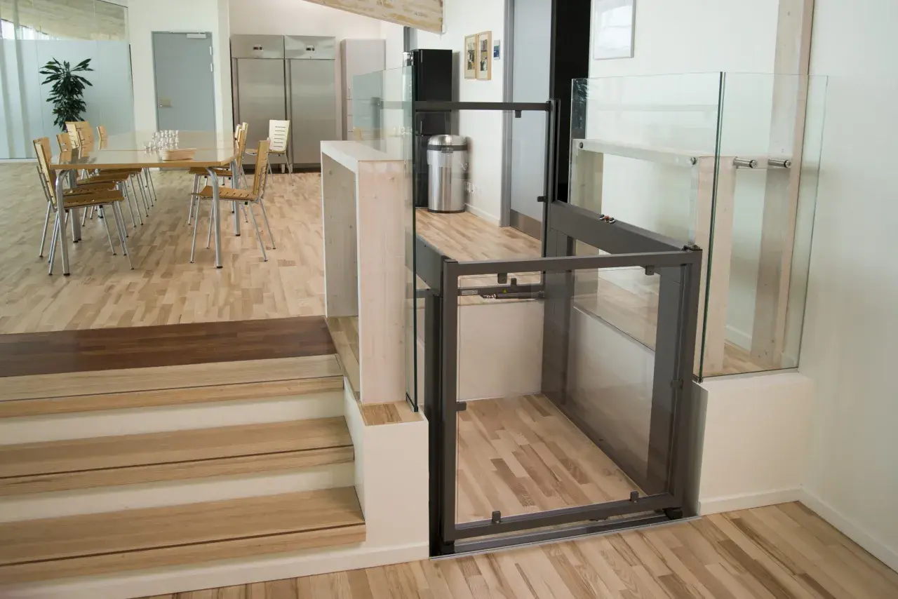 Piattaforma elevatrice per interni con cabina in vetro e struttura metallica, integrata in un ambiente moderno con scale in legno e sala da pranzo.