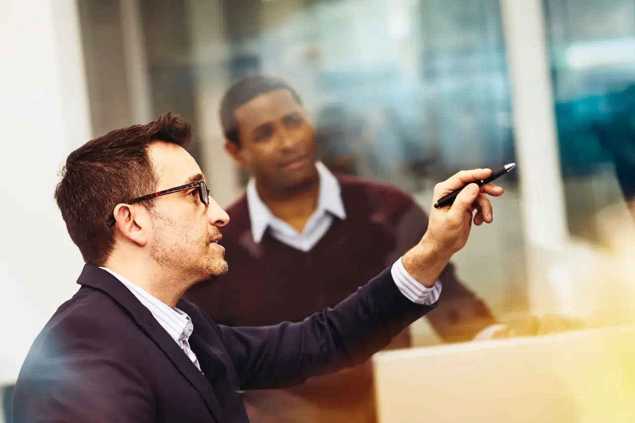 Uomo con occhiali indica un punto su uno schermo, discutendo un **salary review** con un collega in background.