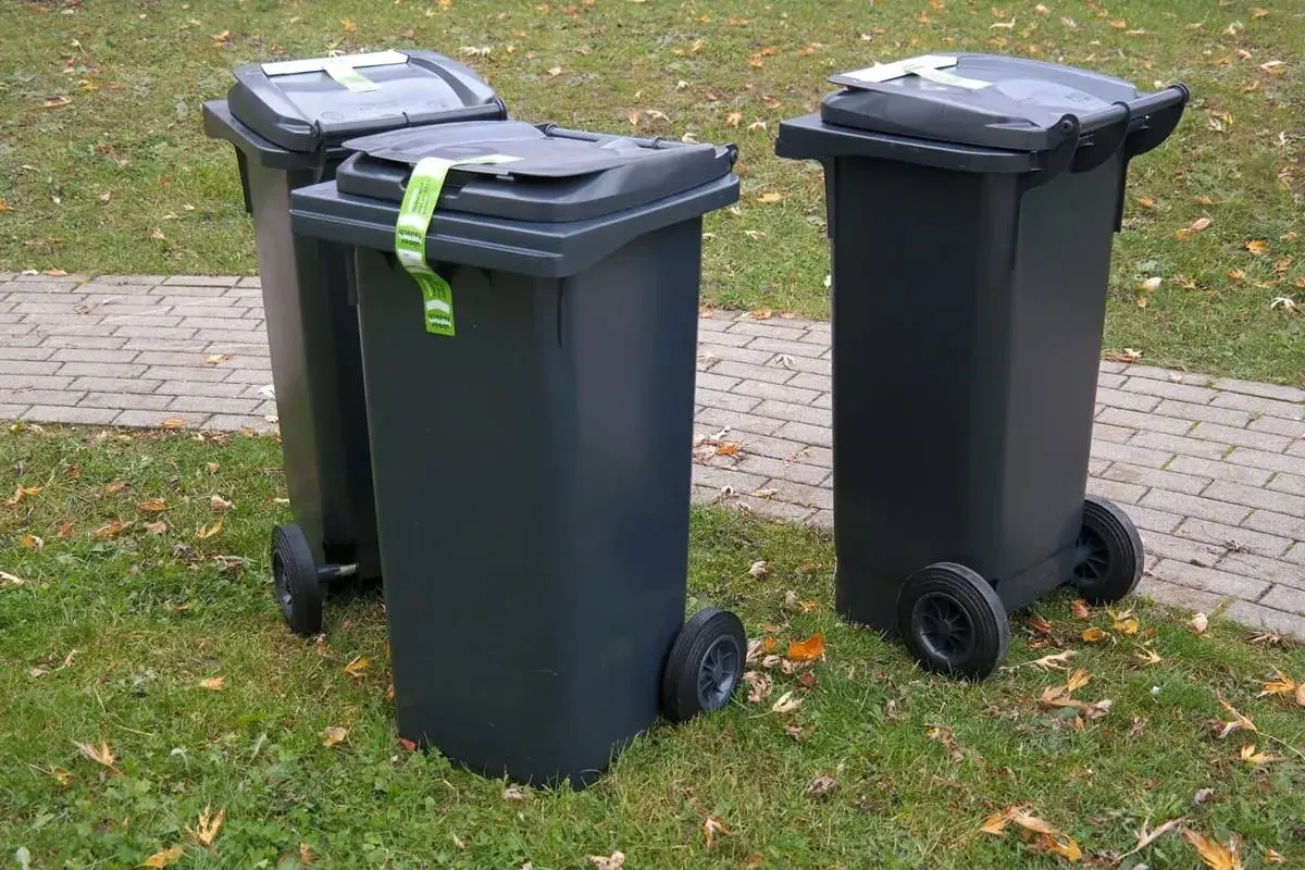 Trzy szare kosze na śmieci stoją na trawie. Zastanawiasz się, gdzie wyrzucić kołdrę z pierza? Sprawdź lokalne przepisy dotyczące odpadów wielkogabarytowych.