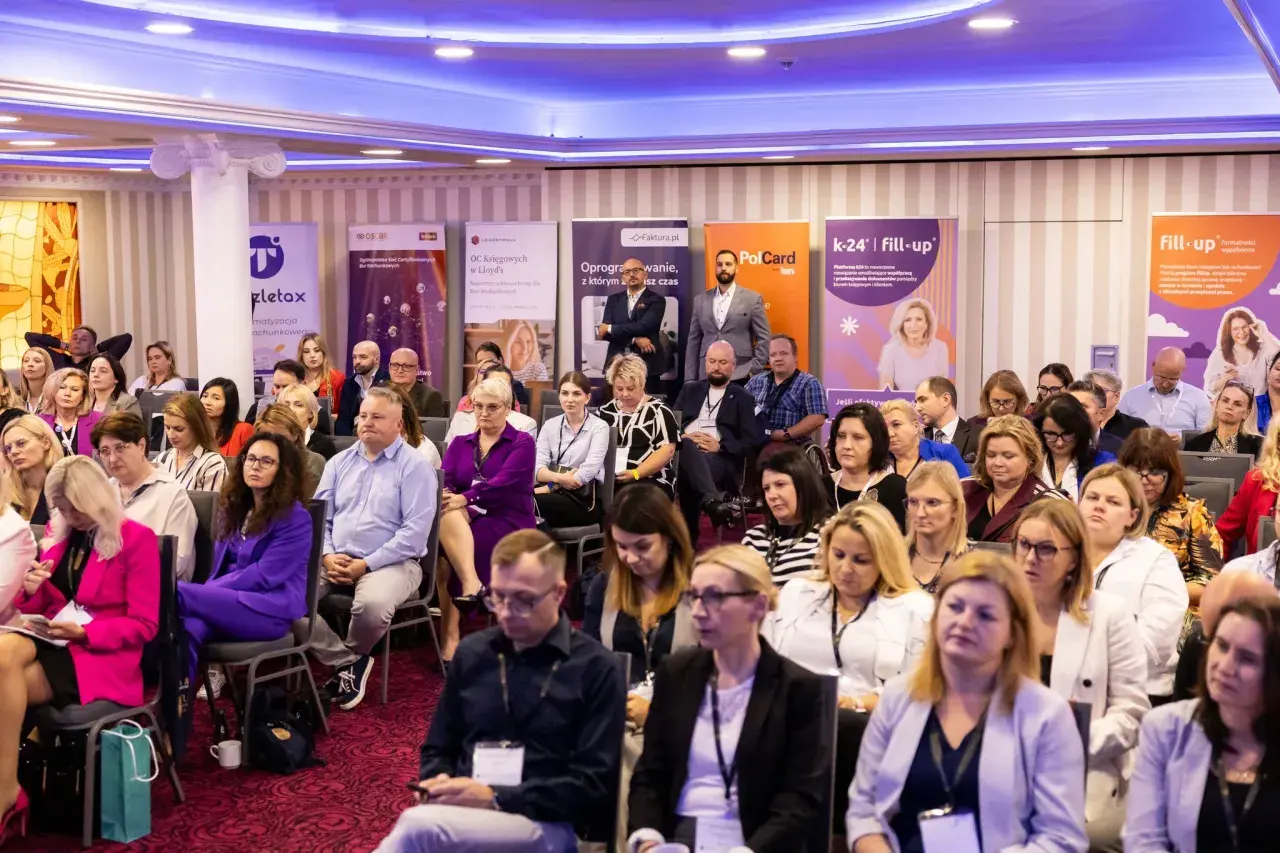 Sala konferencyjna pełna ludzi siedzących w rzędach, z banerami sponsorów w tle.