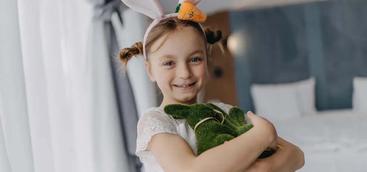 Uśmiechnięta dziewczynka w króliczych uszkach przytula zielonego zajączka. Wesoła wielkanoc w hotelu.