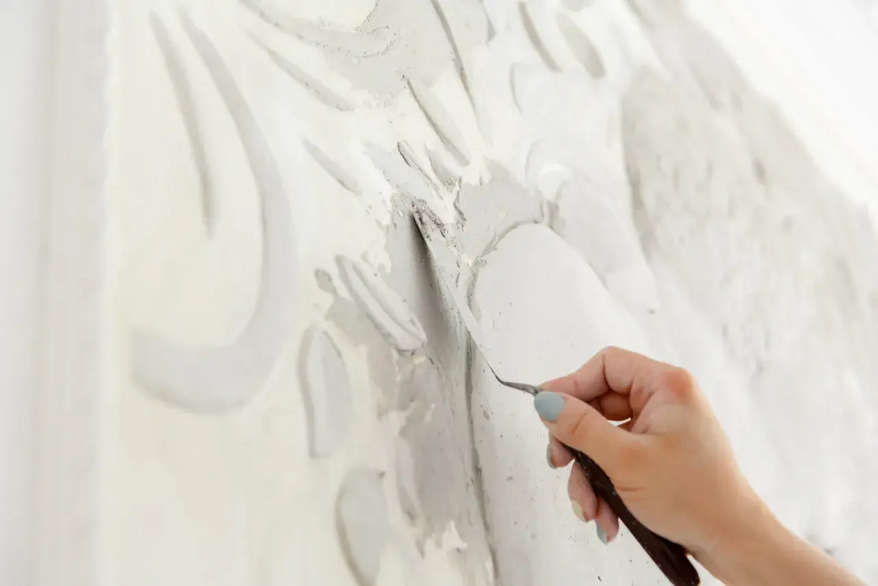 Mano che lavora con uno strumento su uno stucco murale in rilievo, creando un'opera d'arte tridimensionale.