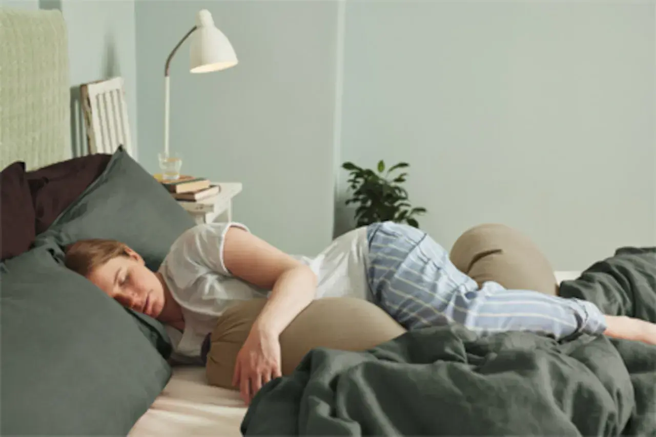 Kvinna sover i sängen, omgiven av kuddar. Hon har på sig randiga pyjamasbyxor. Kanske hon känner av slempropp eller flytning.