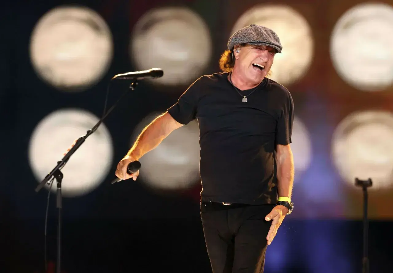 Brian Johnson w czapce i czarnej koszulce śpiewa do mikrofonu na scenie, rozbawiony.