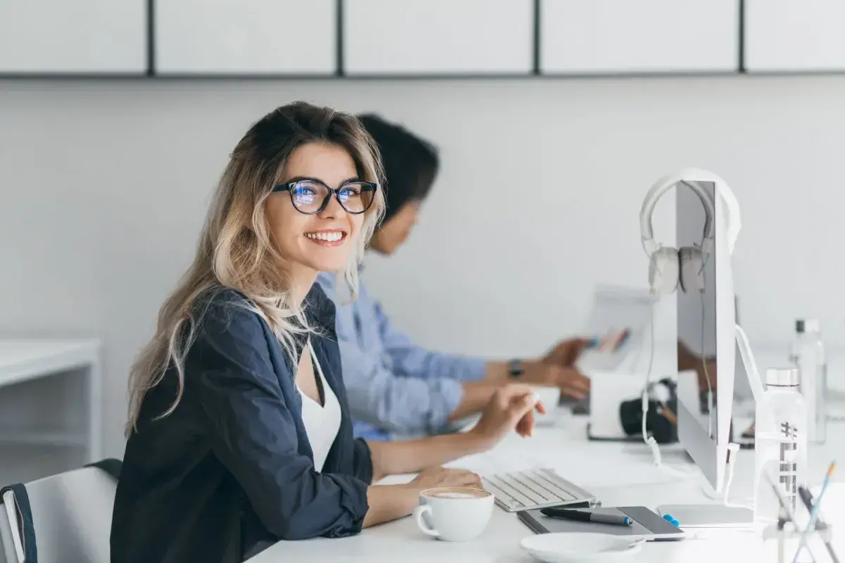 Uśmiechnięta kobieta w okularach pracuje przy komputerze. W tle koleżanka. To obrazek idealnie oddający ducha dyrektywy work life balance.
