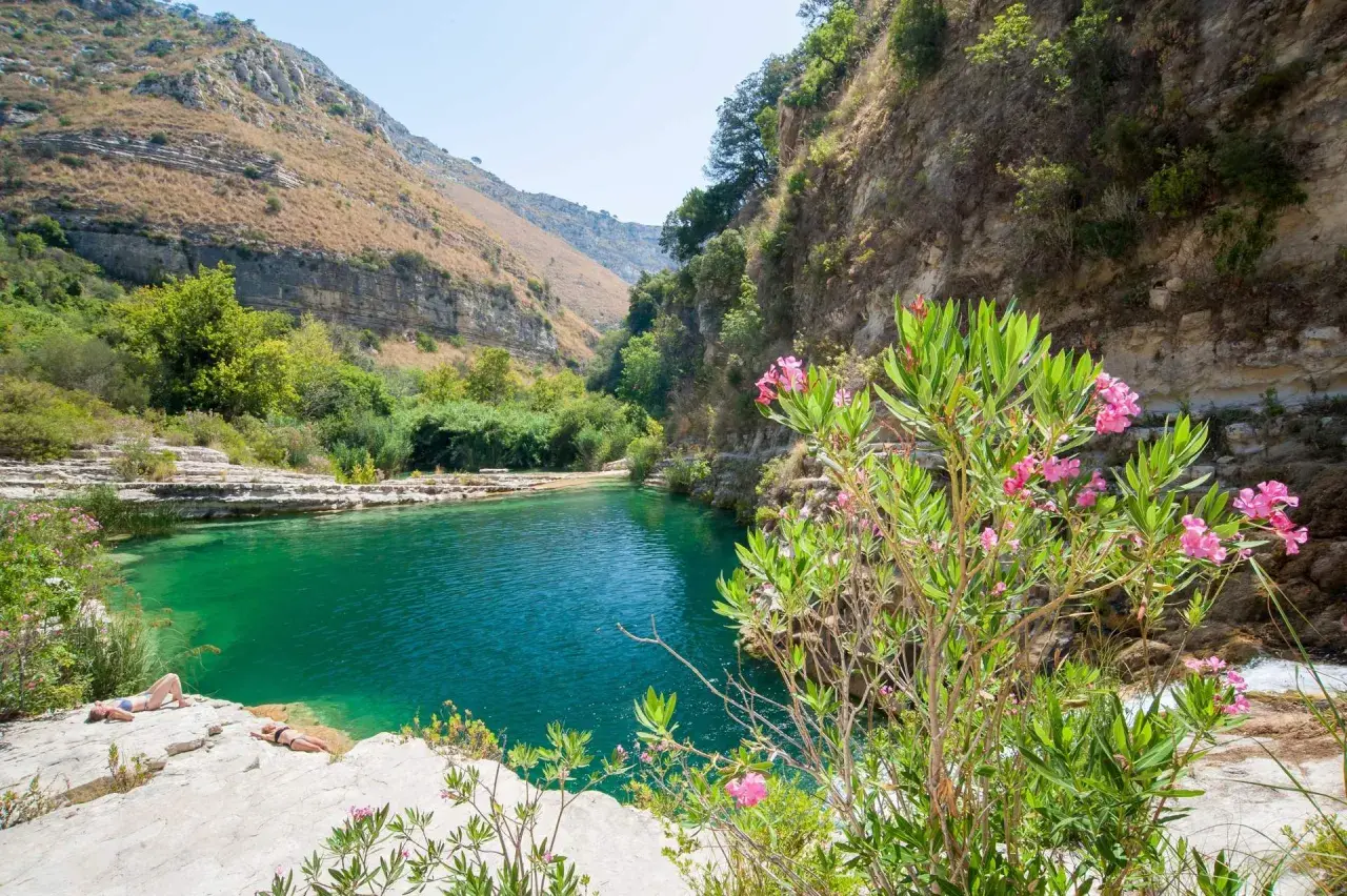 A serene pool of turquoise water reflects the rugged beauty of Sicily nature. Two sunbathers relax on the rocky shore, surrounded by lush greenery and blooming pink oleander flowers.