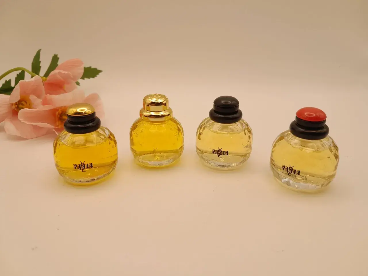 Cztery miniaturowe flakoniki perfum YSL Paris, obok delikatnych różowych kwiatów.