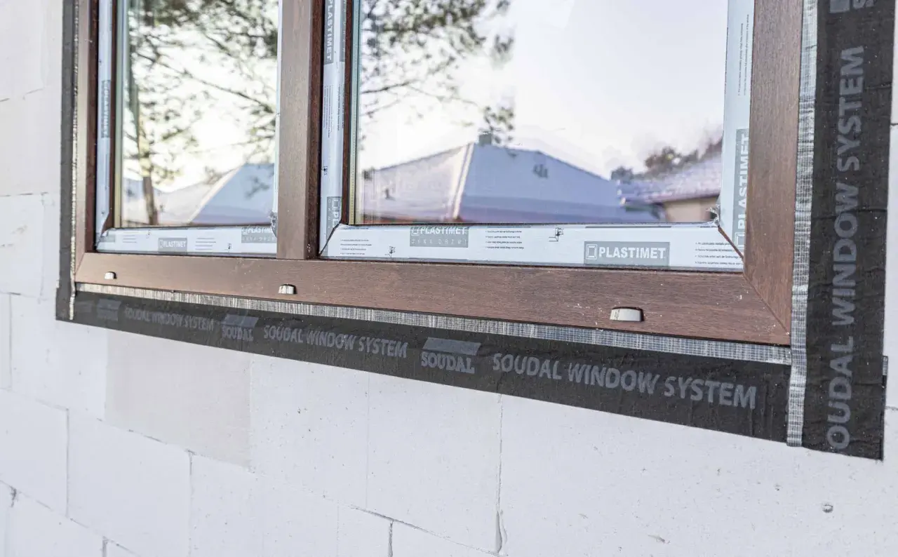 Brązowe okno w trakcie ciepłego montażu z użyciem taśmy SOUDAL WINDOW SYSTEM. Widoczna biała ściana z bloczków.