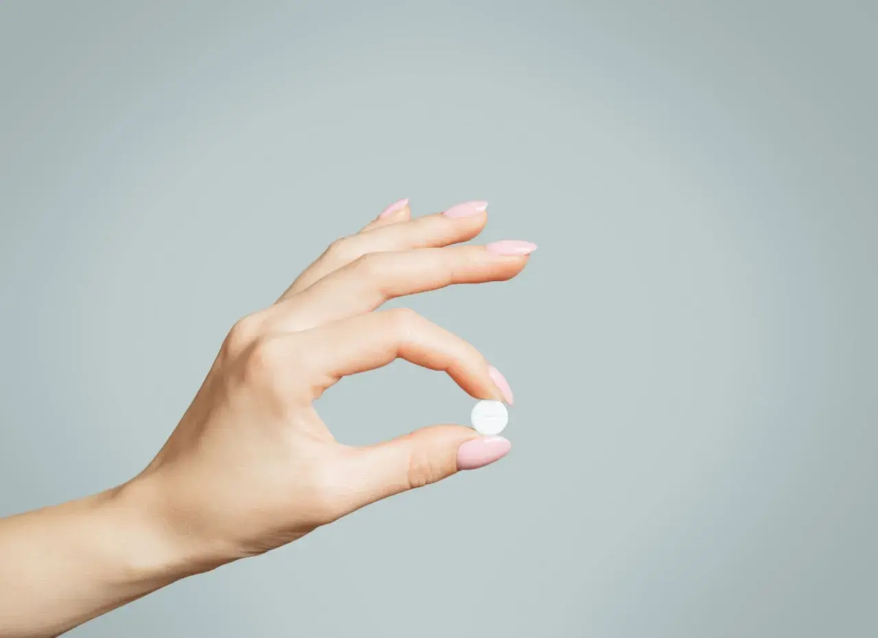 Dłoń z różowymi paznokciami trzyma białą tabletkę, być może zolpidem, na jasnoszarym tle.