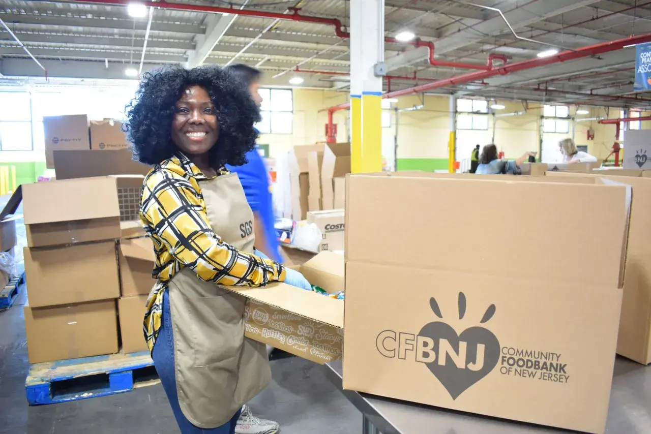 A smiling woman in an apron and gloves packs boxes at a food bank, embodying the spirit of a food bank volunteer job description.